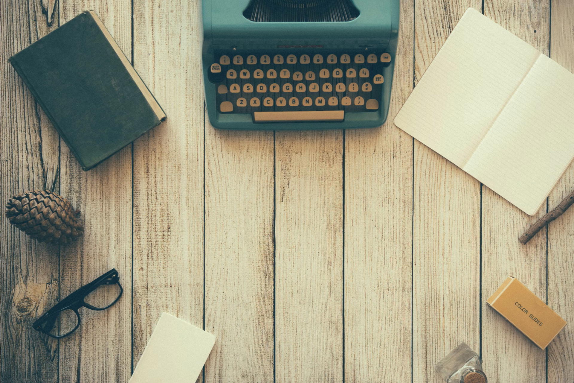 A wooden table with a typewriter, glasses, notebook, and pen placed on top.